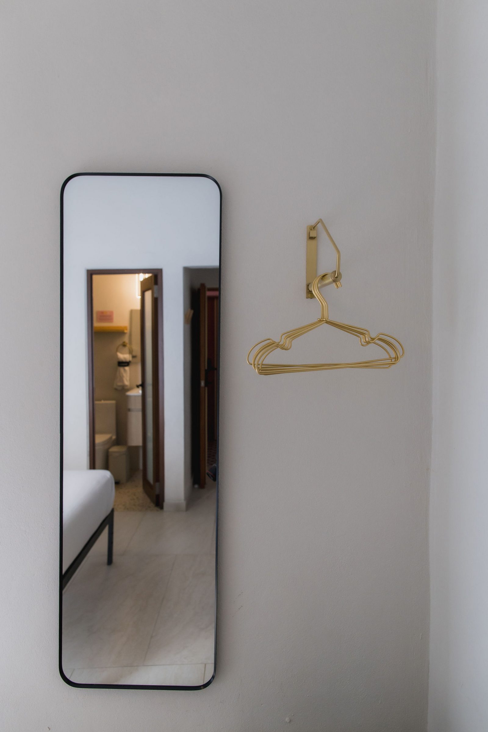 Hotel Luz guest room with full-length mirror and brass wall hooks, bathroom visible beyond